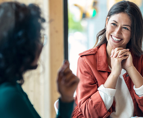 two women engaging in conversation with expressive hand gestures while another person holds a coffee cup in a bright setting